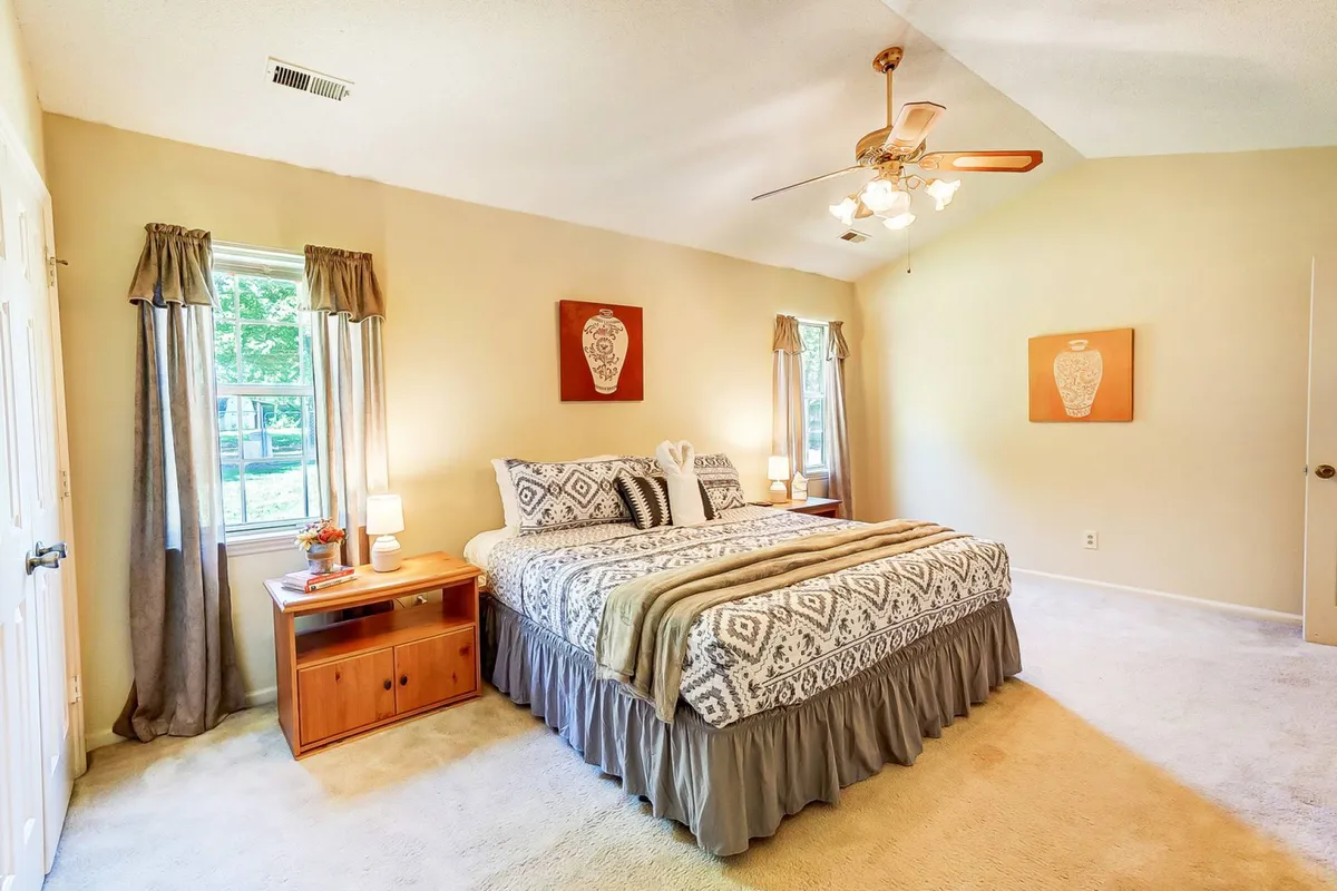 A serene bedroom with a full-size bed dressed in patterned bedding, a wooden nightstand with a lamp, windows with flowing curtains letting in natural light, and decorative wall art. The space exudes calm and comfort, ideal for restful nights.