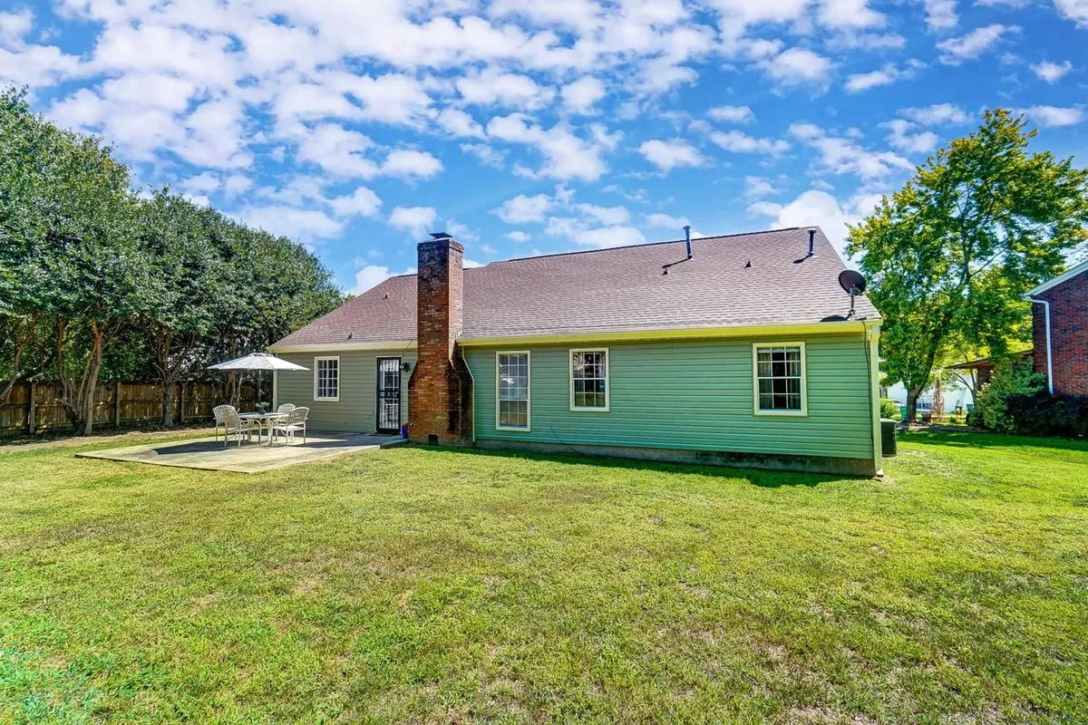 This image features the green exterior of the house, a prominent chimney, a well-maintained lawn, and an outdoor seating area with an umbrella. The area looks welcoming and perfect for outdoor gatherings, with ample space for pets or children to play in the yard.