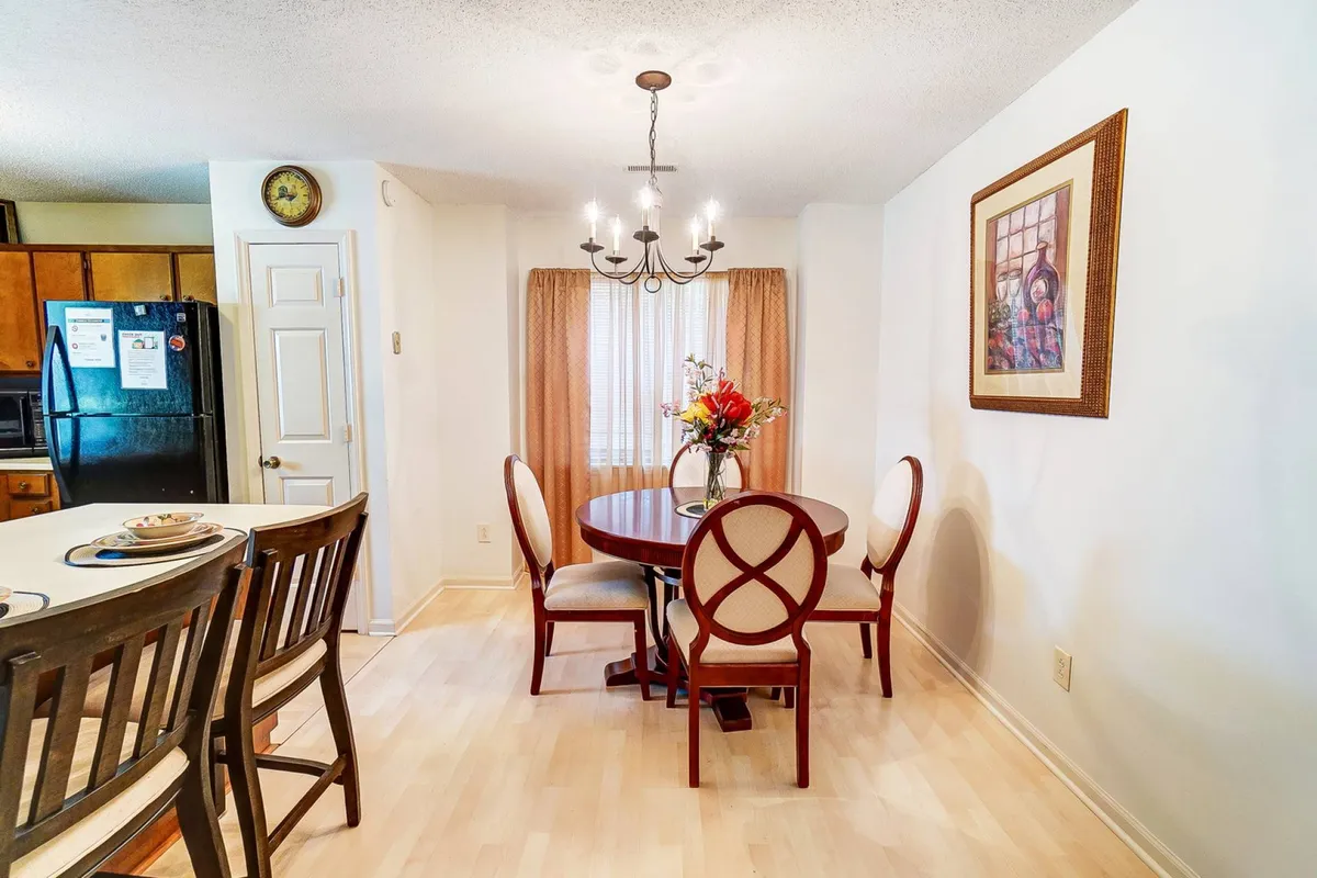 A warmly lit dining area featuring a round table set beneath a chandelier, complemented by a vibrant flower vase and tasteful wall artwork. The space feels inviting—a natural hub for meals and socializing.