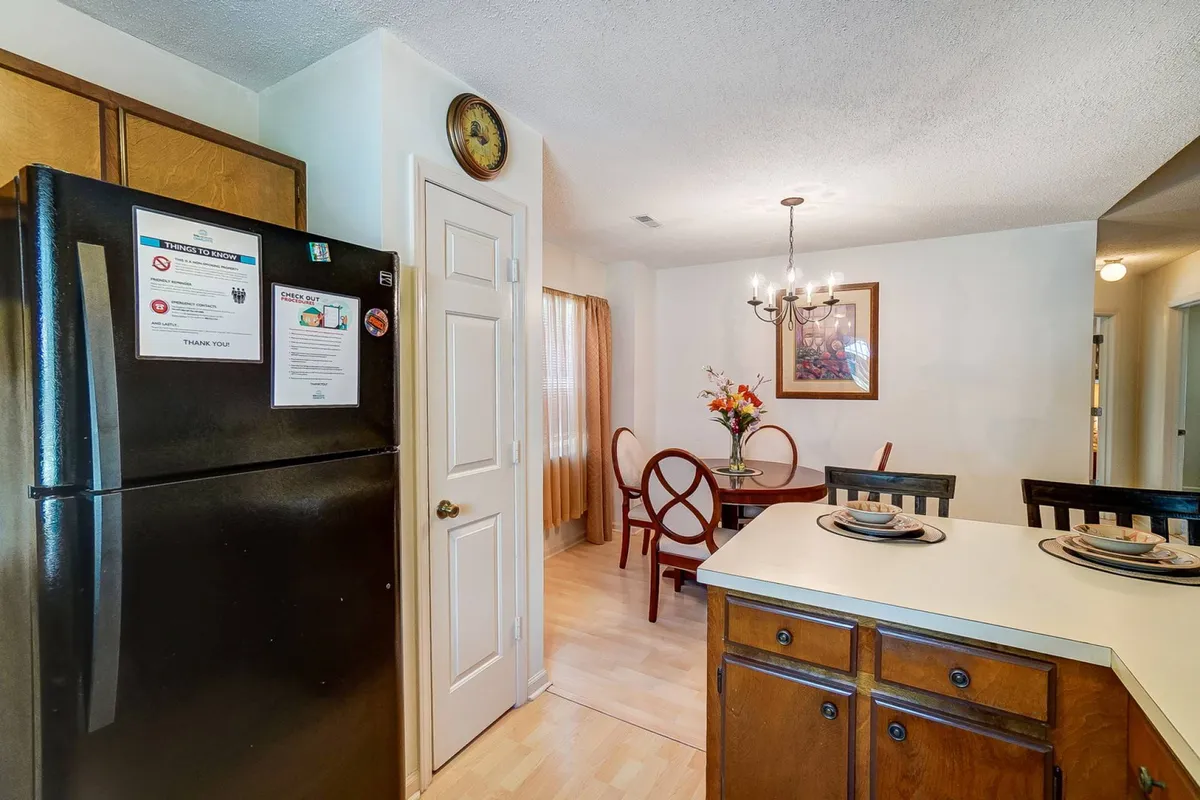 A spacious dining area adjacent to the kitchen, with a breakfast bar, dining table with chairs, chandelier overhead, and a black refrigerator. The space feels open and ideal for casual meals, quick breakfasts, or working remotely in a comfortable setting.