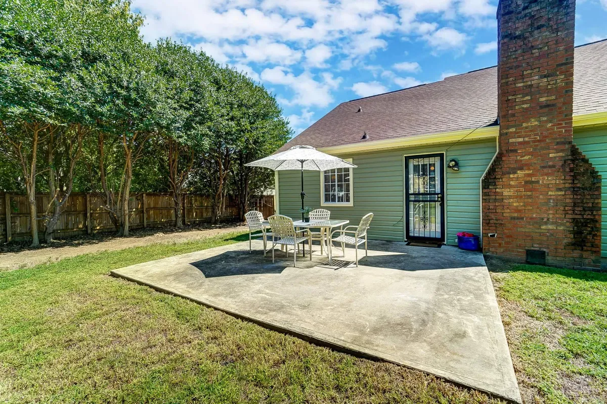 The photo highlights an outdoor table with an umbrella set on a patio, the green siding of the house, a brick chimney, and a landscaped yard. The space is inviting and set up for outdoor dining or morning coffee, emphasizing comfortable seating for four.