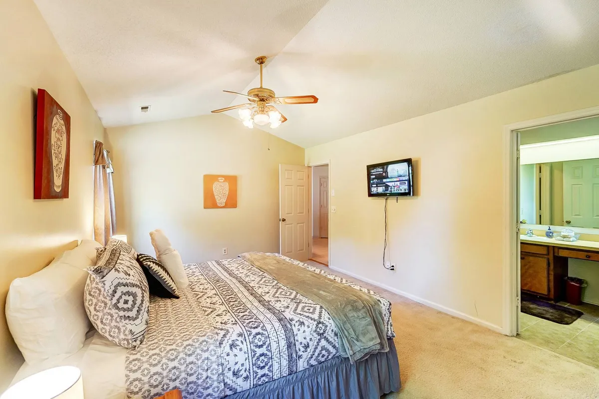 A spacious bedroom featuring a king-size bed with plush bedding, decorative wall art above the headboard, a flatscreen television opposite the bed, and a visible entrance to the ensuite bathroom. Ambient lighting and soft morning sunlight contribute to a relaxing atmosphere.