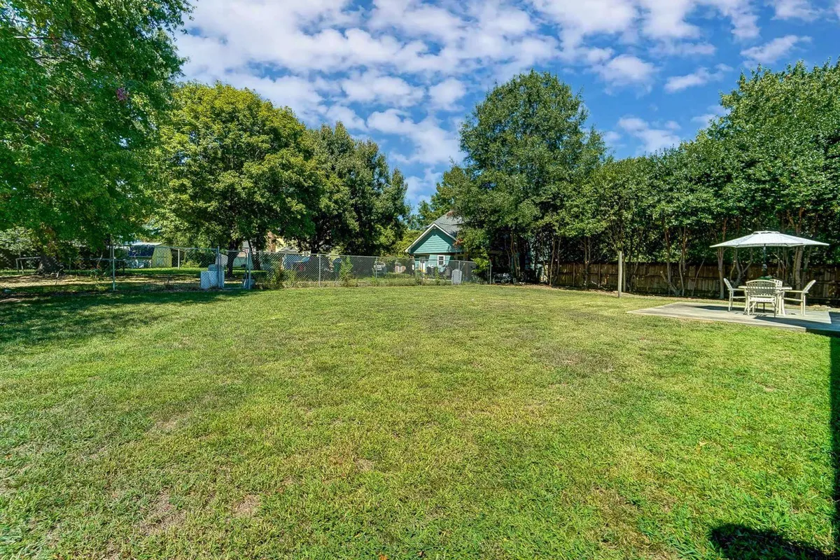 An inviting exterior shot featuring an expansive green lawn, shaded seating area, and a fenced perimeter surrounded by mature trees. The scene suggests a peaceful, private outdoor space ideal for relaxation or small social gatherings before or after attending city events.
