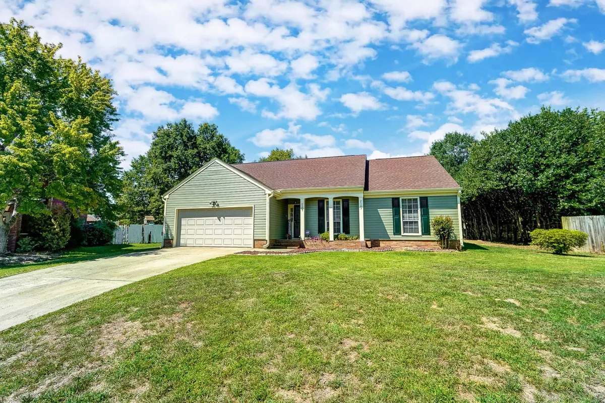 This image displays the front view of the house with its green siding, a welcoming front porch, well-maintained lawn, driveway, and tall trees in the background. The spacious driveway is visible, providing easy access and parking for guests.