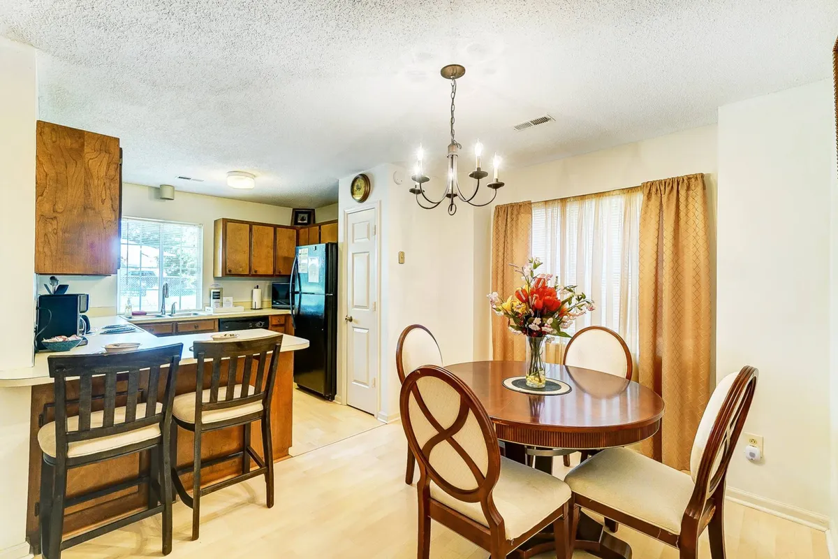 A warm, welcoming dining area with a round table adorned with a floral centerpiece, an elegant chandelier overhead, and a view into the kitchen with wooden cabinetry. The space invites togetherness and shared meals.