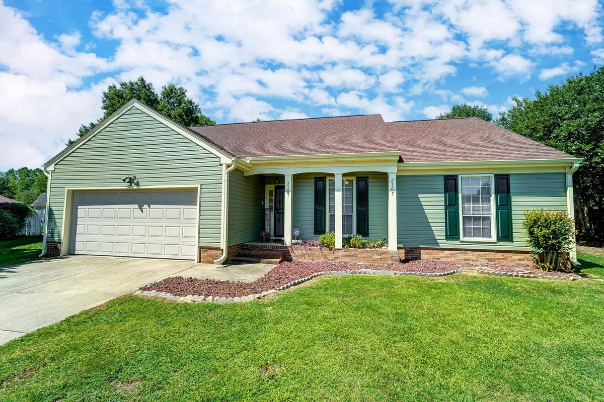 The image features the charming exterior of a single-family home with green siding, a sturdy brick foundation, and decorative shutters. A welcoming front porch suggests a perfect spot for relaxing, with plenty of space for morning coffees or watching the sunset. The inviting landscaping hints at outdoor activities like gardening or gatherings with friends and family.